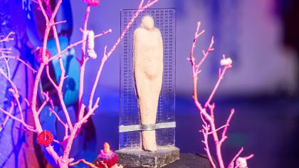 Die BBA-Statue im Detail: Eine steinerne Figur umgeben von einer transparenten Tafel mit aufgedrucktem Binärcode. Die Statue wird umrahmt von Zweigen in rosa.