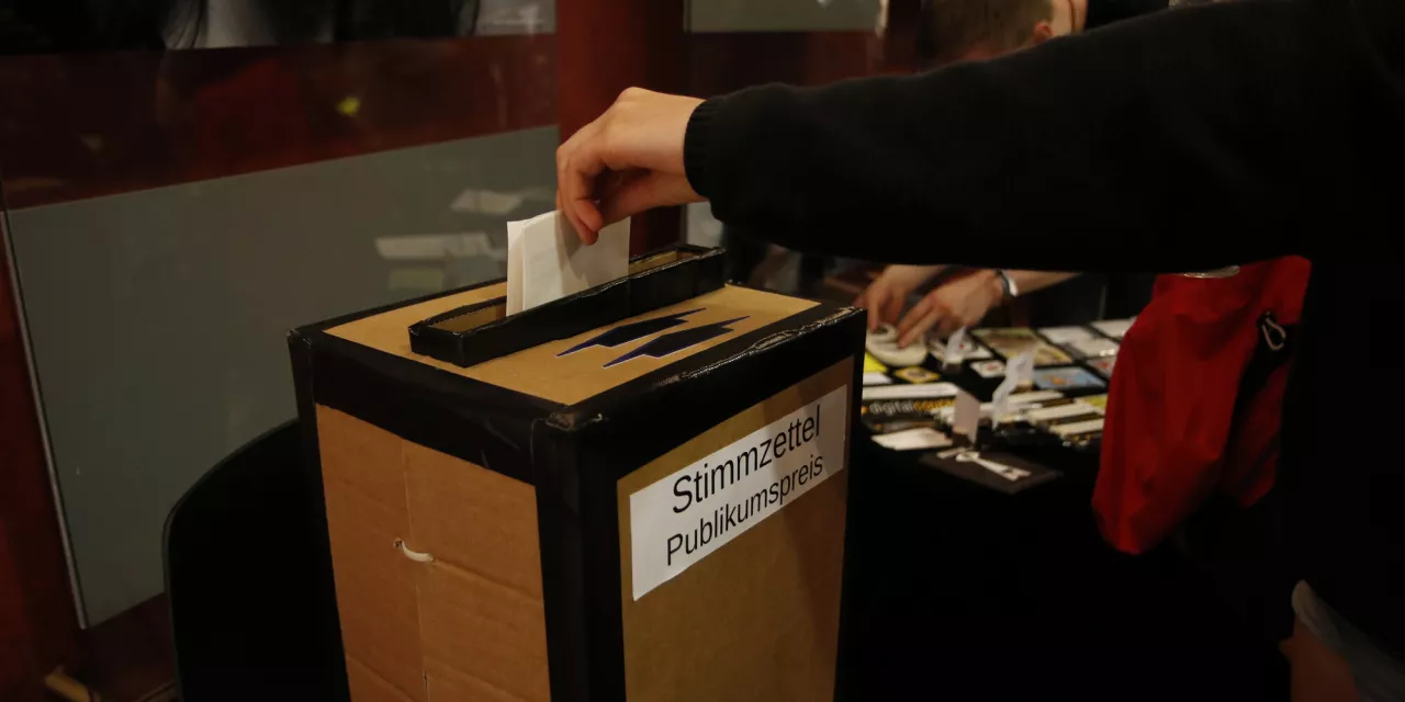 Ein Stimmzettel zum Publikumspreis wird in eine Wahlurne gesteckt.