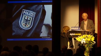 Rolf Gössner am Redner.innenpult. Im Hintergrund das Wappen der Polizei auf einem Jackenärmel (Detailaufnahme).