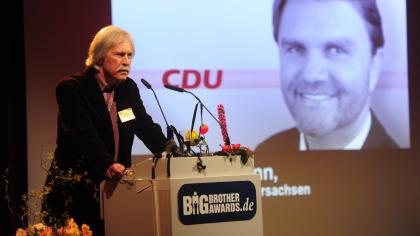 Rolf Gössner am Redner.innenpult. Im Hintergrund eine Portraitaufnahme von Uwe Schünemann und das Logo der CDU.