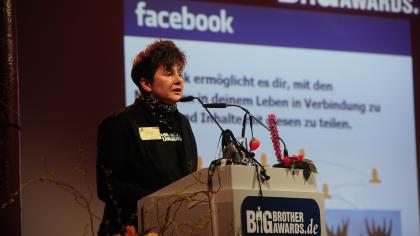 Rena Tangens am Redner.innenpult der BigBrotherAwards 2011. Im Hintergrund ein Screenshot der Startseite von Facebook.