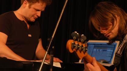 Zwei Musiker (einer neben einem Laptop, der andere mit einem Bass)