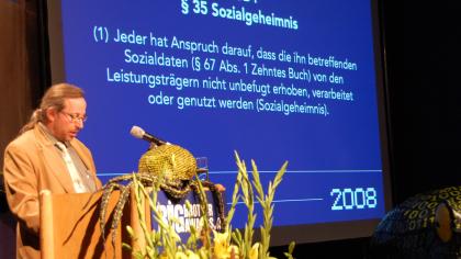 Werner Hülsmann am Redner.innenpult. Im Hintergrund ein textauszug aus dem SGB (weiße Schrift auf blauem Grund).