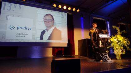 padeluun am Redner.innenpult. Im Hintergrund das Prudsys-Logo sowie ein Portraitfoto von Jens Scholz (Vorstand).