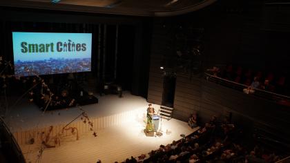 Rena Tangens am Redner.innenpult aus der Ferne. Im Hintergrund eine Stadt, darüber der Schriftzug "Smart Cities".