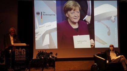 Rolf Gössner am Redner.innenpult. Im Hintergrund ein Foto von Angela Merkel.