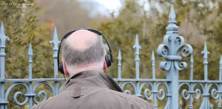 Person von hinten, die Kopfhörer trägt und vor einem Zaun steht.