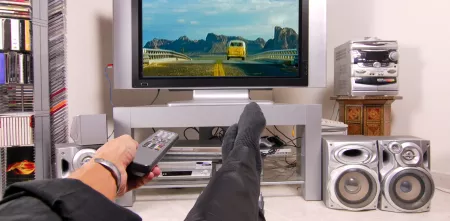 Point of view: Eine Person, die vor einem Fernseher sitzt. Die Fernbedienung in der Hand zeigt Richtung Fernseher. Die Füße sind hochgelegt.