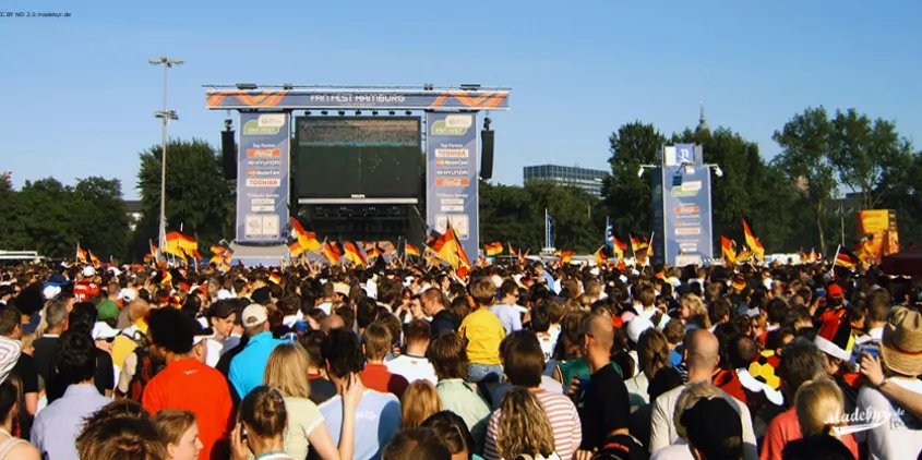 Blick auf eine Menge Fußballfans mit Deutschlandfahnen vor einer grißen Leinwand.