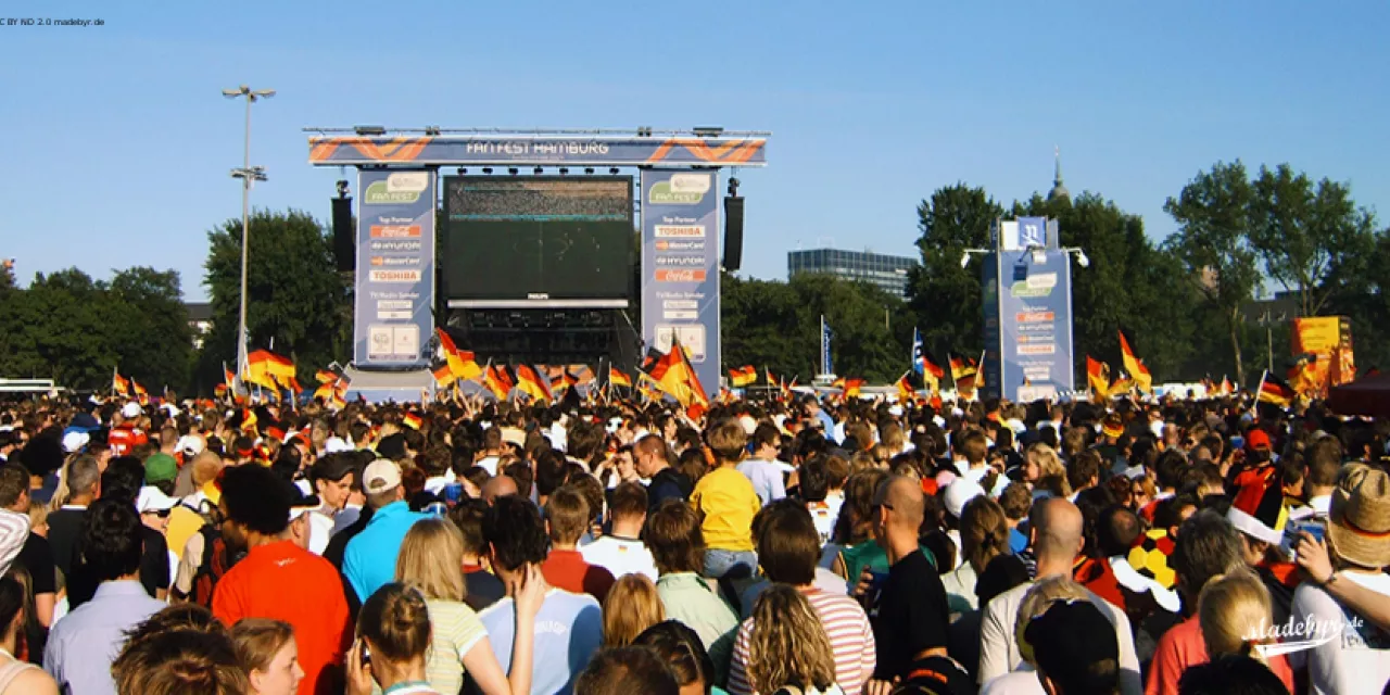 Blick auf eine Menge Fußballfans mit Deutschlandfahnen vor einer grißen Leinwand.