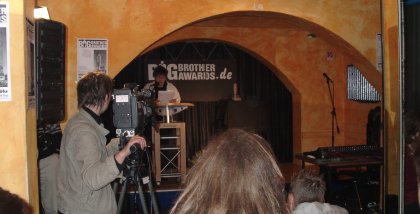 Rena Tangens am Redner.innenpult (aus der Ferne; im Vordergrund eine Kamera).