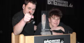 padeluun und Rena Tangens lehnen auf dem Redner.innenpult. padeluun zegt mit einem Finger Richtung Kamera. Der BigBrotherAward steht zwischen ihnen.