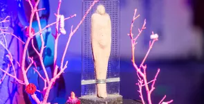Die BBA-Statue im Detail: Eine steinerne Figur umgeben von einer transparenten Tafel mit aufgedrucktem Binärcode. Die Statue wird umrahmt von Zweigen in rosa.