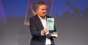 Moderator Anderas Liebold hält ein Buch Richtung Kamera.