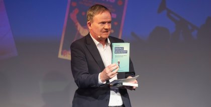 Moderator Anderas Liebold hält ein Buch Richtung Kamera.