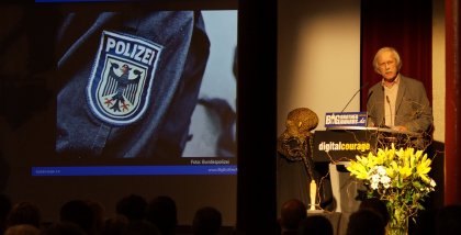 Rolf Gössner am Redner.innenpult. Im Hintergrund das Wappen der Polizei auf einem Jackenärmel (Detailaufnahme).