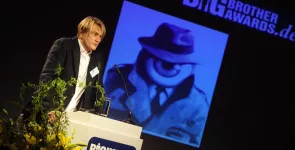 Frank Rosengart am Redner.innenpult. Im Hintergrund eine Collage von einer Person in Mantel und Hut, aber der Kopf ist ein großes Auge.