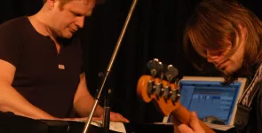 Zwei Musiker (einer neben einem Laptop, der andere mit einem Bass)