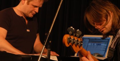 Zwei Musiker (einer neben einem Laptop, der andere mit einem Bass)