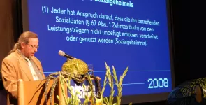 Werner Hülsmann am Redner.innenpult. Im Hintergrund ein textauszug aus dem SGB (weiße Schrift auf blauem Grund).