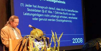 Werner Hülsmann am Redner.innenpult. Im Hintergrund ein textauszug aus dem SGB (weiße Schrift auf blauem Grund).