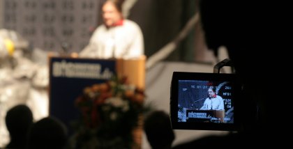Alvar Freude am Redner.innenpult beobachtet durch eine Filmkamera.