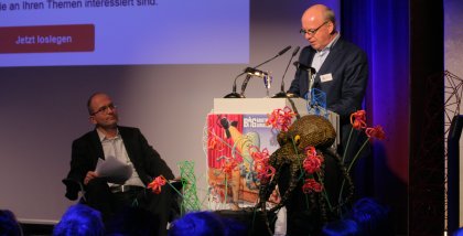 Peter Wedde am Redner.innenpult. Neben ihm sitzt Sönke Hilbrans auf einem Stuhl.