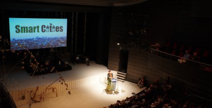 Rena Tangens am Redner.innenpult aus der Ferne. Im Hintergrund eine Stadt, darüber der Schriftzug "Smart Cities".