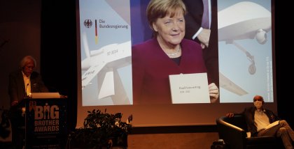 Rolf Gössner am Redner.innenpult. Im Hintergrund ein Foto von Angela Merkel.